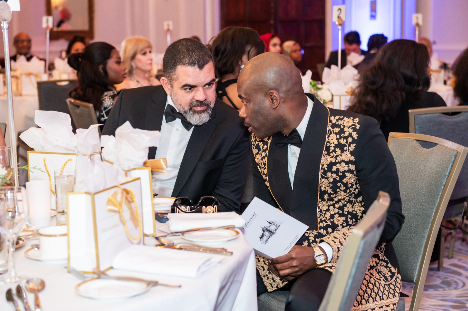 Two guests in tuxedos sharing a private exchange at the gala dinner table
