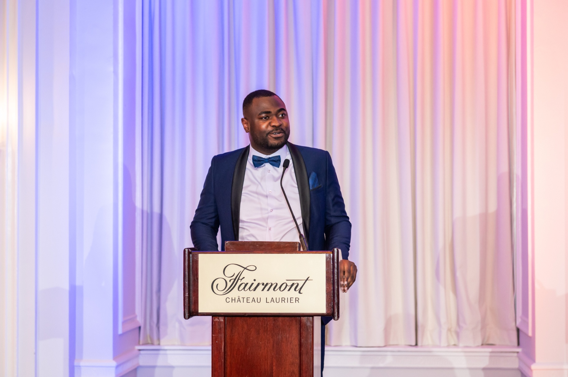Gala emcee in navy tuxedo speaking from the Fairmont Château Laurier podium